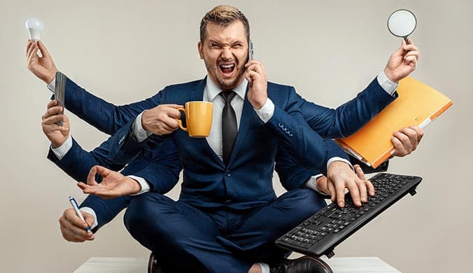 man in suit sitting on the desk holding phone, laptop, pen, lightbult, coffee mug and more indicating multitasking