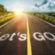 A scenic road with the words ‘let’s go’ painted on the asphalt against a blue sky with the sun on the horizon