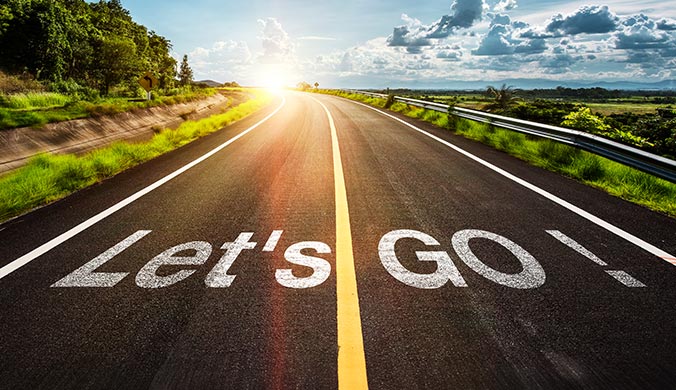 A scenic road with the words ‘let’s go’ painted on the asphalt against a blue sky with the sun on the horizon