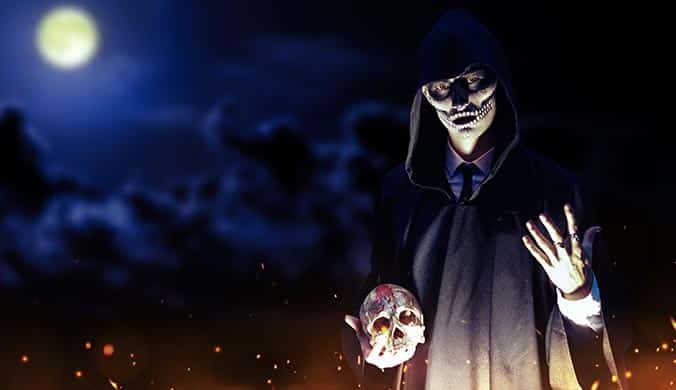 An image of a cloaked and masked man standing by a fire holding a skull during full moon