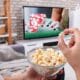 A close-up photo of a man holding a bowl of popcorn watching television at home