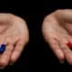 A photo of female hands holding a red and blue pill in each hand, on a dark background